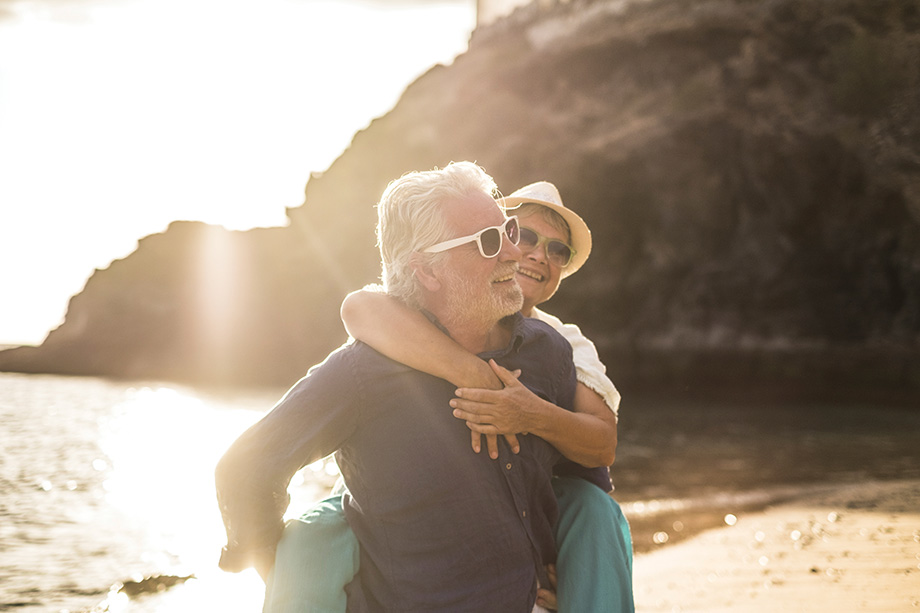 adults caucasian people couple in love with man carrying the woman and both smile and laugh a lot - sunset and backlight at the beach - vacation lifestyle married forever concept for happy matures retired
