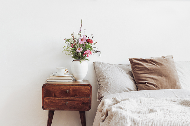 Cup of coffee and books on retro wooden bedside table. Rustic white ceramic vase with bouquet of pink cocmos and zinnia flowers. Beige linen and velvet pillows in bed, Scandinavian interior, bedroom.