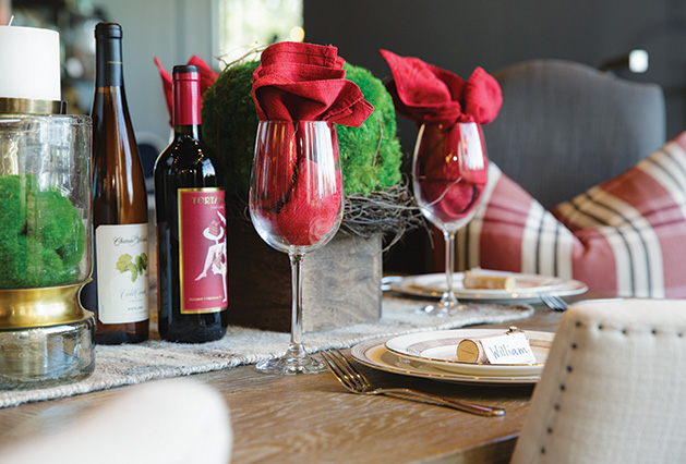 A holiday table spread featuring wine and wine glasses, candles, a place setting and more.