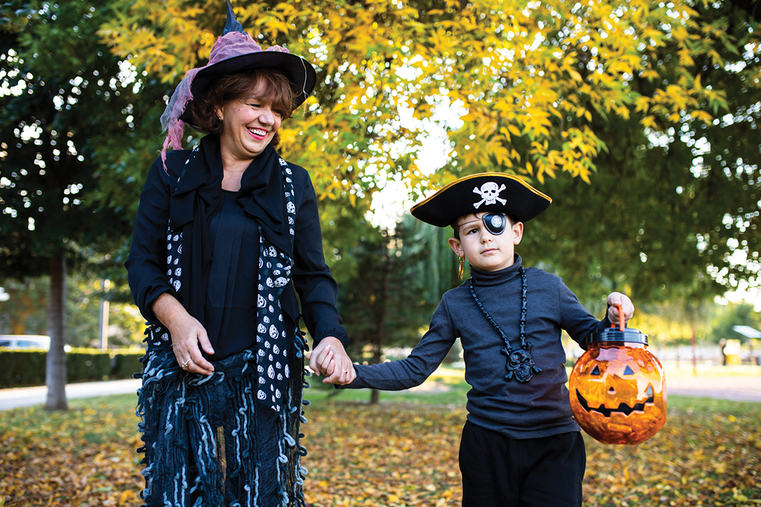 A mature woman dressed as a witch and a cute boy in a pirate costume are holding hands and going trick-or-treating